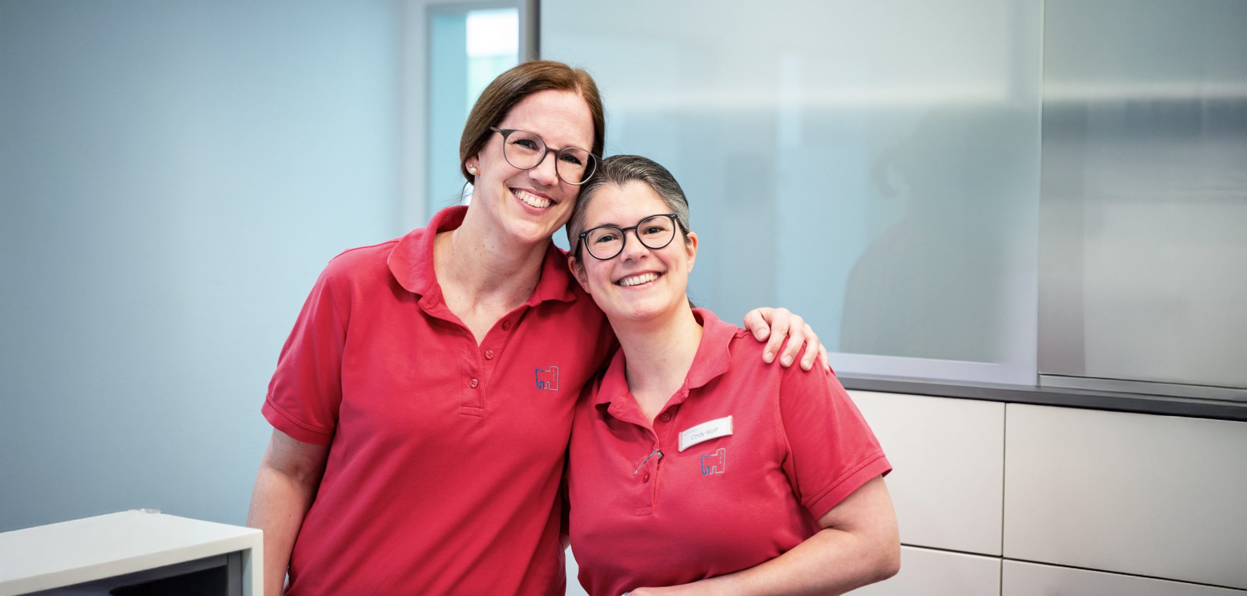 Zwei Mitarbeiterinnen mit roten Poloshirts halten sich im Arm in der Praxis Güntzer und Brandstätter